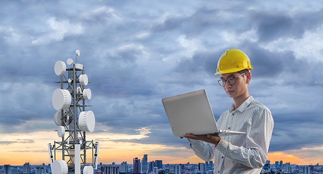 Engineer in protective helmet conducting a signal test from a tower, holding a laptop in his hands.
