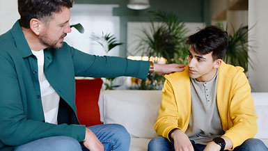 Man in green blazer comforts young man in yellow hoodie on white couch.