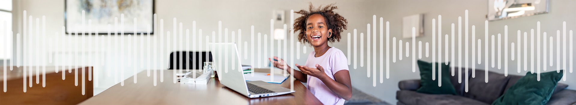 A little girl in front of a laptop, listening through a set of black earbuds, smiling
