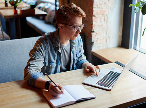 Young man in glasses working on a laptop while taking notes in a cozy café with natural light.