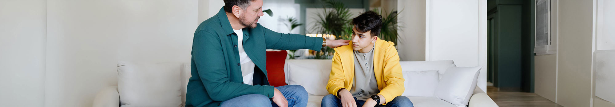 Man in green blazer comforts young man in yellow hoodie on white couch.