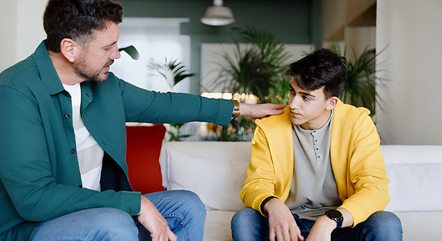 Man in green blazer comforts young man in yellow hoodie on white couch.