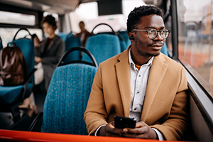 A man sits on a bus while holding a phone and wearing wireless earbuds