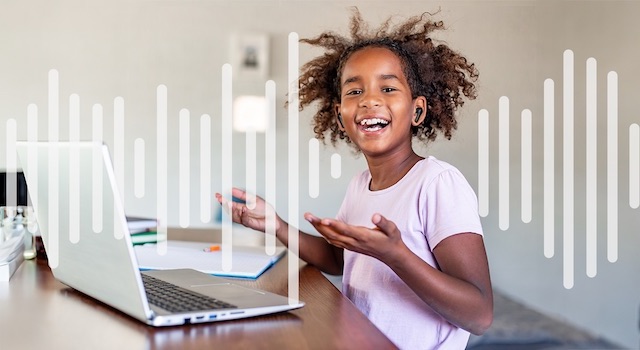 A little girl in front of a laptop, listening through a set of black earbuds, smiling