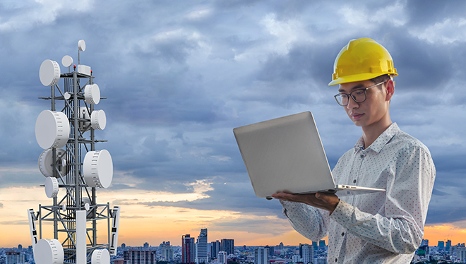 Engineer in protective helmet conducting a signal test from a tower, holding a laptop in his hands.