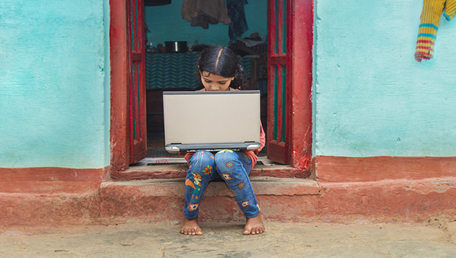 Child sitting on a step in a doorway holding an outdated laptop in a small village in India.