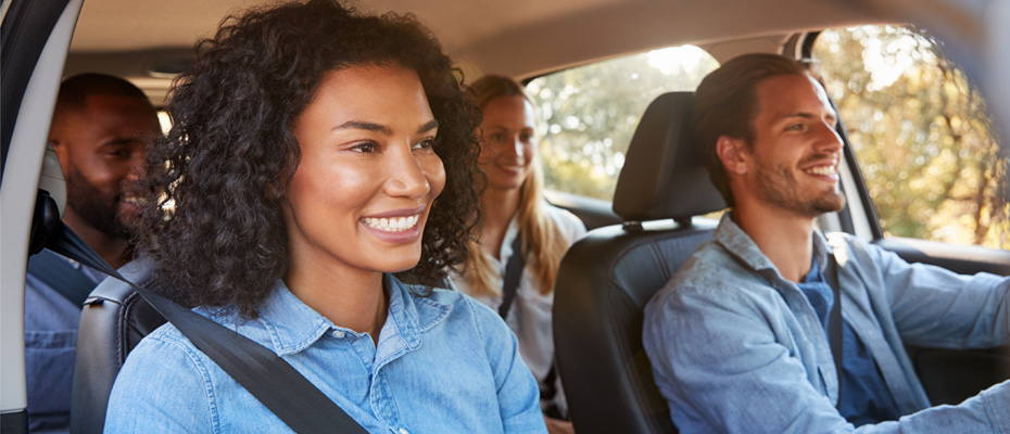 Image shows front view of four smiling people of difference races and genders inside interior of a car while commuting to work.