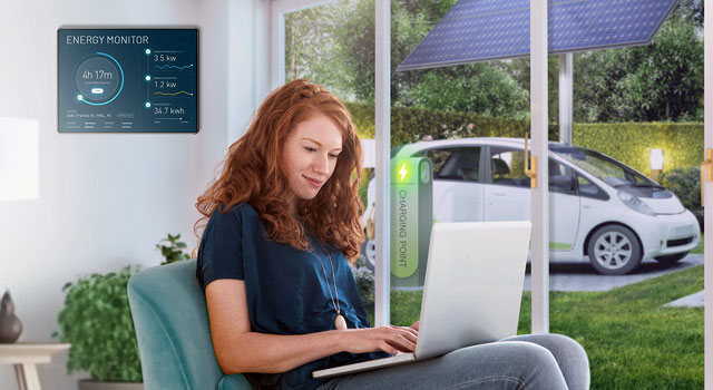 A girl in her living room with a wall-mounted energy monitor, overlooking an EV charging station and solar panels outside.