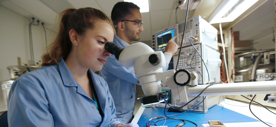 Image shows engineers and data scientist; one female and one male working in a lab. One is on a microscope machine and the other is pushing buttons on data machines.