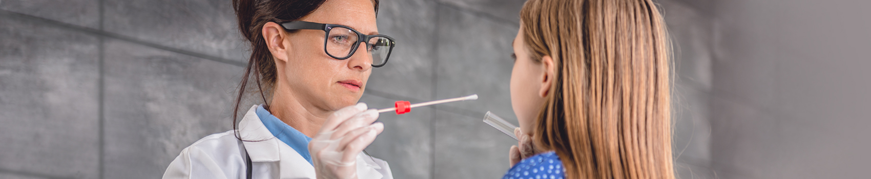 Image shows a female family doctor in a Doctor's office setting. She is using a test swab and taking a sample from a child's mouth.