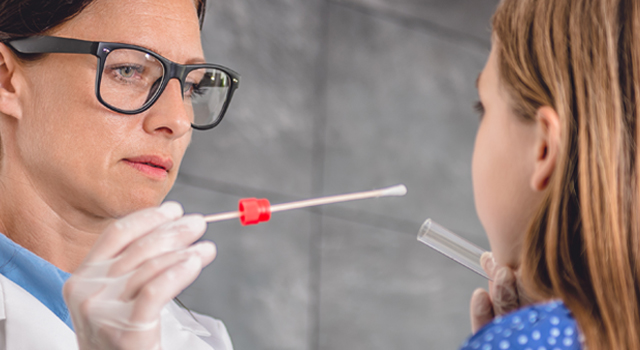 Image shows a female family doctor in a Doctor's office setting. She is using a test swab and taking a sample from a child's mouth.