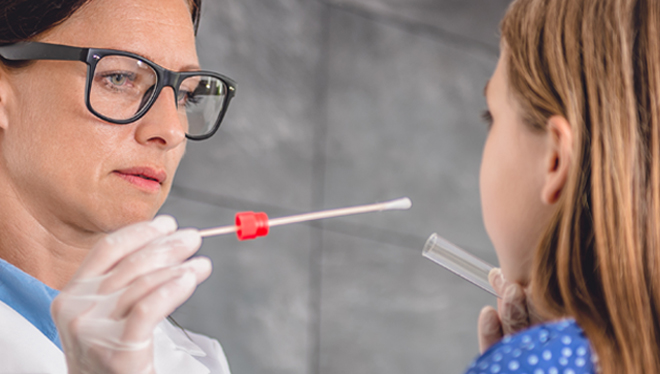 Image shows a female family doctor in a Doctor's office setting. She is using a test swab and taking a sample from a child's mouth.